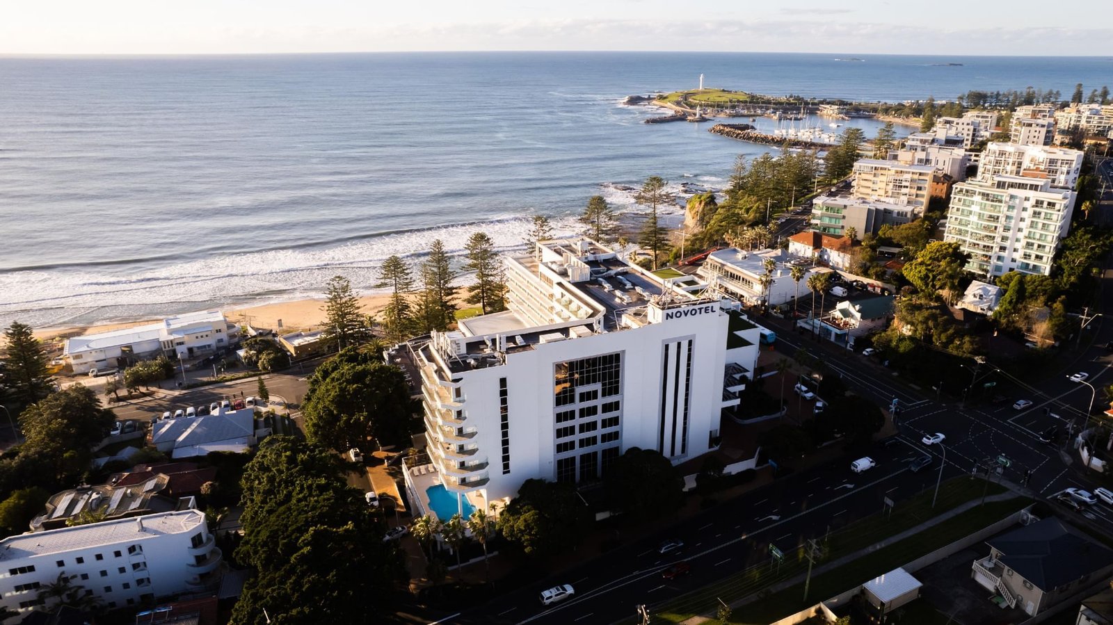 Aerial view of the Novotel Wollongong Northbeach among the Norfolk pines beside North Wollongong Beach, with the lighthouse on the headland beyond